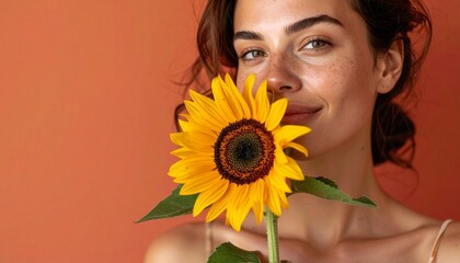 ピーチ色のマット背景に、そばかすが特徴的な白人女性が鮮やかな黄色のひまわりを抱え、ミニマルなアクセサリーと落ち着いた表情で横を見つめる静かなポートレート。

