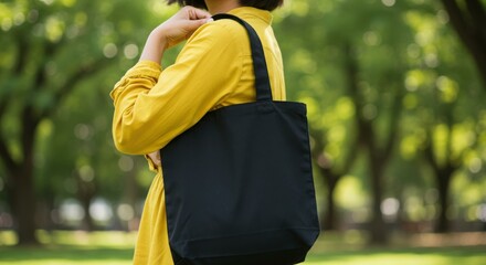 Naklejka premium Woman in yellow shirt carrying a black tote bag over her shoulder in a park setting.