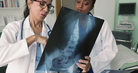 Senior female radiologist giving consultation to young doctor about mri scan shot of spine and pelvic bones, explaining examination result. Diverse medical colleagues discussing trauma, diagnosis - Powered by Adobe