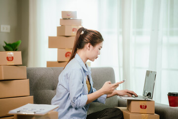 Young Asian businesswoman happily packing e-commerce orders in home office with laptop, cardboard boxes, and wireless technology for online retail.