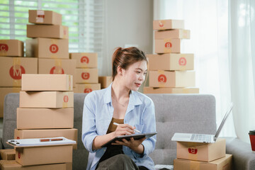 Young Asian businesswoman happily packing e-commerce orders in home office with laptop, cardboard boxes, and wireless technology for online retail.