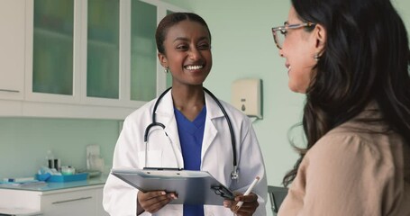 Positive young African doctor filling patients records form on meeting, talking to senior woman with friendly smile, asking about health complaints, therapy, treatment, writing notes - Powered by Adobe