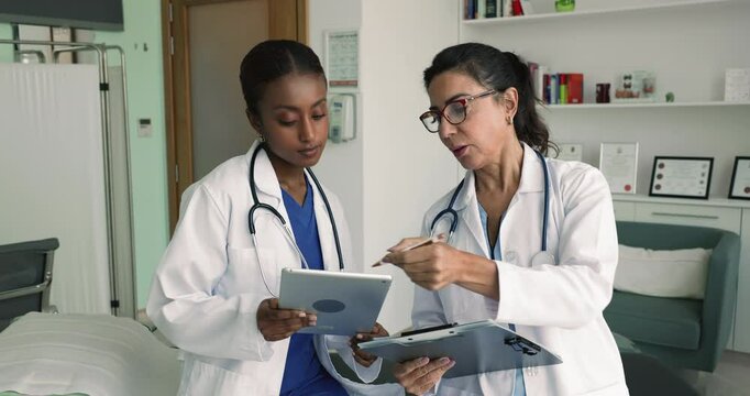 Mature mentor doctor training young African intern, explaining medical records, electronic data on tablet. Two diverse practitioners comparing data on tablet and paper documents, discussing report