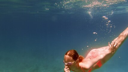 Young girl gliding underwater in blue ocean, wearing pink bikini and goggles, exploring marine environment with graceful movements during summer vacation - Powered by Adobe