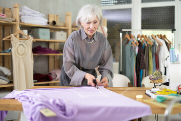 Mature dressmaker stands in the workshop and cuts fabric. Fabric cutter prepares material before...
