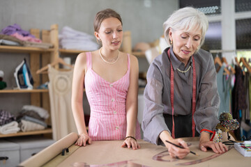 Female dressmakers stand in a sewing workshop, making a sketch on paper and discussing a new...