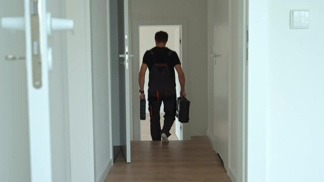 Professional repairman in overalls walks down hallway carrying toolboxes in both hands. Worker leaving bright modern interior after work. Concept of home repair or renovation