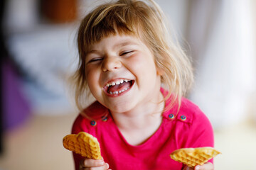 Portrait of happy little preschool girl holding fresh baked heart waffle. Smiling hungry toddler child with sweet biscuit wafer. Sweet sugar belgian waffles.