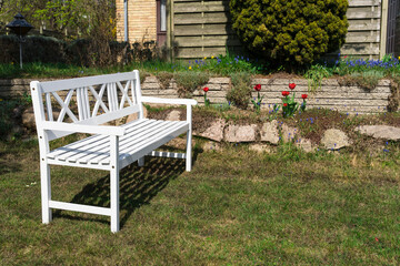 A white, three-seater garden bench with armrests sits on green grass in front of a stone wall with flowers 