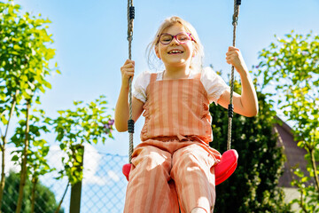 Happy little school girl with eyeglasses having fun on a swing in the garden. Smiling and laughing...