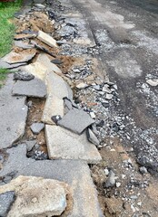 Damaged road surface with broken asphalt and gravel. Uneven pavement and debris scattered along the roadside. Signs of construction or repair work in progress.