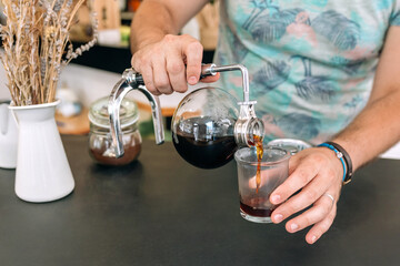 Close up of unrecognizable barista expertly pours brewed coffee from Japanese siphon coffee maker into a glass, showcasing the art of specialty coffee preparation.