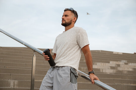 Sportsman resting on a railing, holding a water bottle, enjoying a well-deserved break after an intense training session on the stairs
