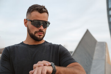 Fototapeta premium Sportsman checking time on his smartwatch while wearing sunglasses, with modern architecture in the background