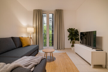 Modern apartment living room with sectional sofa, TV stand, plant and bright balcony window view