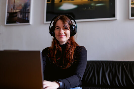 Brunette woman in a home office setting using a laptop and wearing headphones on a couch
