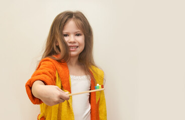 Caucasian young girl in orange bathrobe holding toothbrush with multicolored toothpaste.