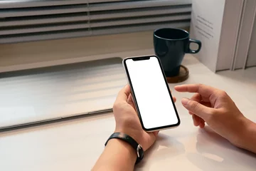 Fotobehang Close up of hands holding smartphone with blank screen at desk with natural light © Prathankarnpap