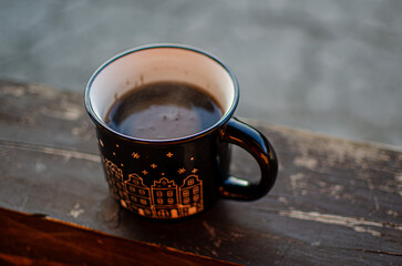 Original black cup with painted houses and coffee. The mug is resting on a beam. A couple made from root in the background of mountains.