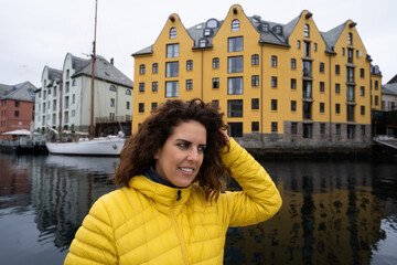 Portrait of young brunette woman looking away touching her hair, wearing a yellow anorak, with the Brosundet Canal in Alesund, Norway, behind her, with art nouveau buildings. Norwegian tourist concept