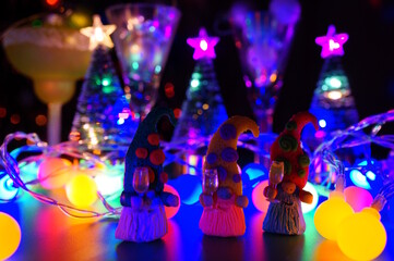 Three toy gnomes made of plasticine with glasses of champagne on the background of a glowing garland.