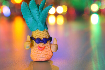 Toy pineapples made of plasticine on the background of a glowing garland. Festive mood.