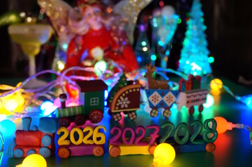 A toy train made of plasticine with the dates 2026, 2027 and 2028 on the background of Christmas decorations.