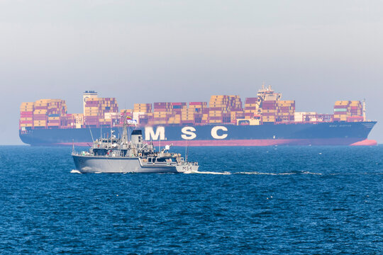 Minehunter vessel M54 sails in front of the massive container ship MSC Sofia on open waters during maritime operations.