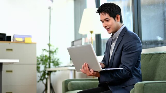 Asian businessman in suit using laptop during remote work meeting. Smiling and confident in modern office