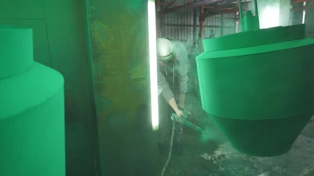 Industrial painting process in chamber at factory. Painter spraying green paint on detail in special booth. Technician in safety wear working at production. Master paint metal in specialised workshop