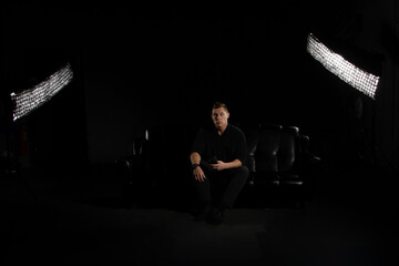 Man Sits on Black Couch Under Studio Lights Holding Phone