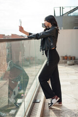 A young, fashionable woman is posing near a glass barrier, looking at herself in a handheld mirror. She is on a rooftop during the daytime, wearing black clothing
