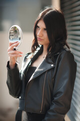 A young woman with dark hair, wearing a stylish black leather jacket, stands outdoors. She holds a round, handheld mirror and attentively examines her reflection
