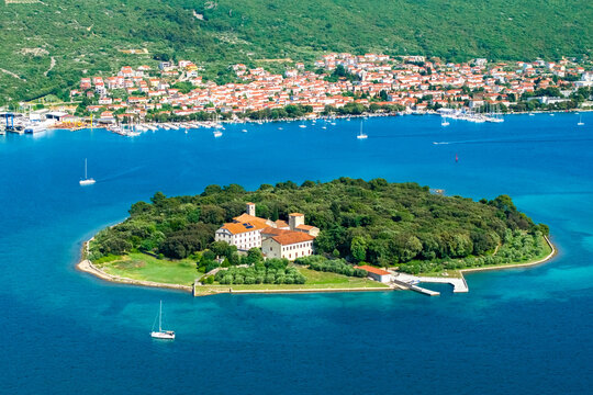 A wide aerial view showcasing Ko&scaron;ljun Island with its monastery, surrounded by calm blue waters and the coastal town of Punat in the background. Sailboats dot the sea