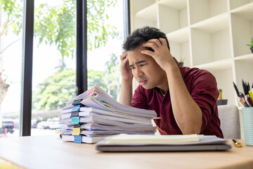 A man in a red shirt is sitting at a desk with a pile of papers in front of him. He is stressed and...
