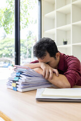 A man in a red shirt is sitting at a desk with a pile of papers in front of him. He is stressed and overwhelmed by the amount of work he has to do
