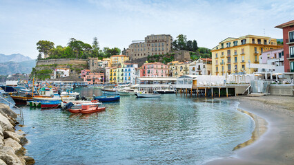 Marina Grande, charming harbor in Sorrento, Italy. Colorful buildings, boats docked in harbor on calm sea water. Hills with lush green trees, cloudy sky create scenic European landscape