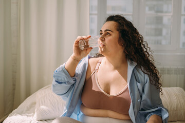 A positive young curly-haired plus-size woman drinks water after waking up in bed.Healthy lifestyle.