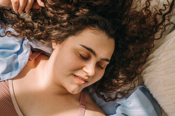 A portrait of a young, attractive plus-size woman with dark curly hair lying in bed with her eyes closed.