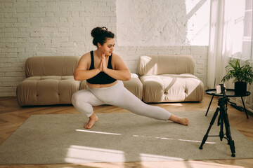 A young curly-haired plus-size woman practices yoga at home, online broadcast.Healthy lifestyle.