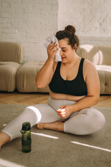 A tired young curly-haired plus-size woman sits on the floor after a workout at home, drinks water and wipes sweat with a towel.Healthy lifestyle.