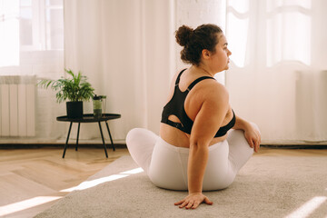 A young curly-haired plus-size woman stretches at home,sits in the lotus position,rear view.Healthy...