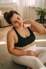 A tired young curly-haired plus-size woman sits on the floor after a workout at home and wipes...
