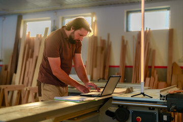 Confident male carpenter using laptop in workshop, designing new furniture, surrounded by wood...
