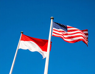 A photo with a bright blue sky background shows two national flags flying on flagpoles. The American flag and the Indonesian flag