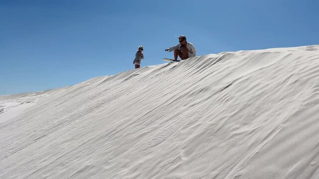 Mother and her daughter enjoy sandboarding on a big dune under a clear blue sky and warm sunlight.