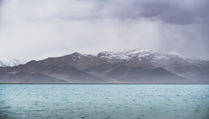 Lake Karakul with blue water and mountain peaks in the background, cloudy weather and sky with...