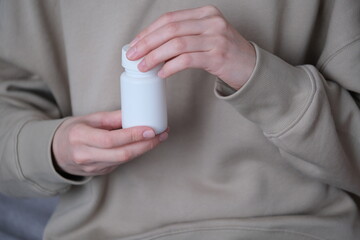 A person gently twists open a white pill bottle using one hand, dressed in a soft beige sweatshirt while seated in a cozy indoor setting.