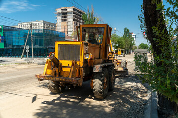 Large yellow motor grader operates on city street, smoothing ground before asphalt application,...