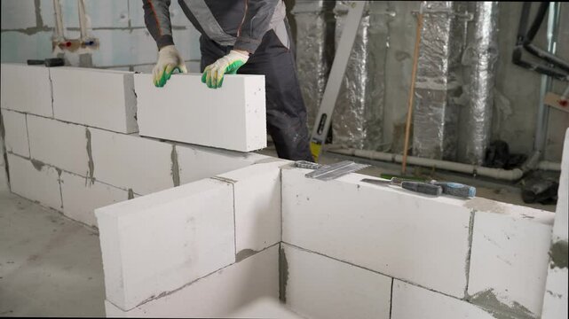 Construction worker carefully placing aerated concrete blocks, building a wall inside a house under construction